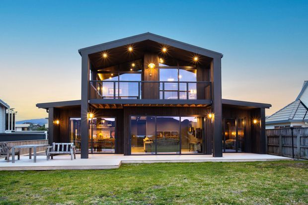 Exterior view of a contemporary two-storey house at dusk, featuring a prominent gabled central volume, dark vertical cladding, extensive glazing, an upper-level balcony, and warm integrated exterior lighting. A lawn and deck/patio occupy the foreground, with neighbouring homes visible at the sides.