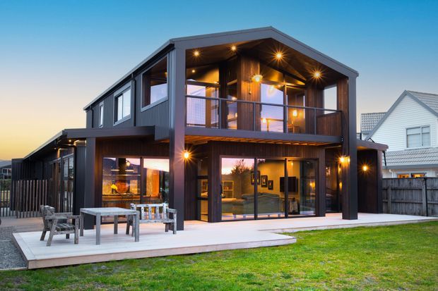 Two-storey contemporary residential house photographed at dusk, featuring dark vertical cladding, a prominent upper-level covered balcony, extensive glazing, exposed timber soffits, warm integrated lighting, and a pale timber deck opening onto a lawn. A neighbouring white gabled house is visible in the background.