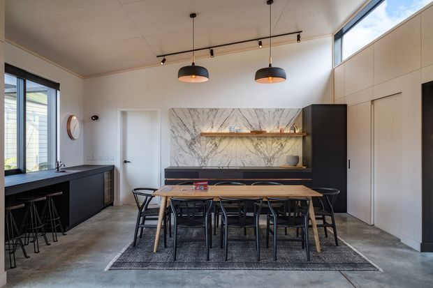 A contemporary open-plan dining and kitchen area with a long timber dining table and black chairs set on a rug. The space features dark kitchen cabinetry, a stone island bench, polished concrete flooring, pale walls and ceiling, a dramatic marble-look feature wall with timber shelving, and a large run of upper glazing that brings in natural light. Black pendant lights and track lighting add contrast.