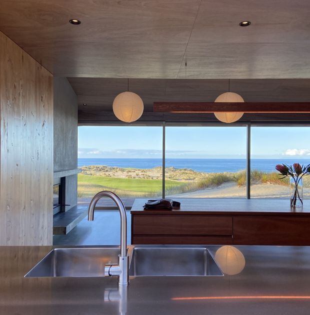 A refined contemporary kitchen with extensive timber wall and ceiling finishes, dark wood cabinetry, a broad stainless-steel island with double undermount sinks, and a sculptural gooseneck tap. Large floor-to-ceiling glazing frames a clear ocean and coastal-dune outlook. Warm spherical pendant lights and integrated recessed lighting create a calm, understated atmosphere.