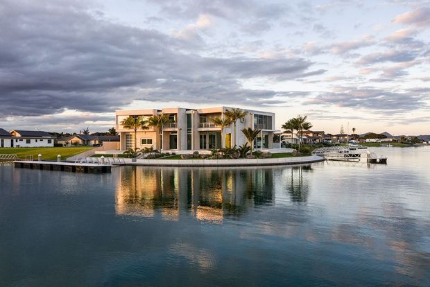 Wide exterior view of a contemporary multi-storey waterfront residence on a narrow landscaped waterfront site, with extensive glazing, balconies, light-coloured rendered walls, palm trees, a private dock and calm reflective water in the foreground. The scene is photographed at dusk beneath a dramatic cloudy sky.