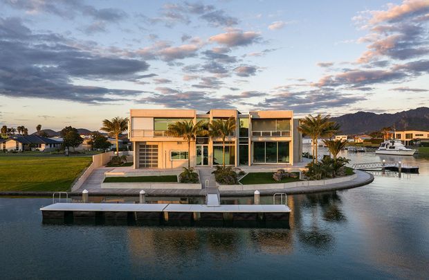 Exterior view of a contemporary multi-storey waterfront residence on a landscaped canal frontage. The home has a rectilinear form, warm light-coloured facade panels, extensive floor-to-ceiling glazing, balconies, a flat roofline, and a private dock with a boat. Palm trees and neighbouring homes frame the scene, with mountains visible in the distance under a dramatic evening sky.