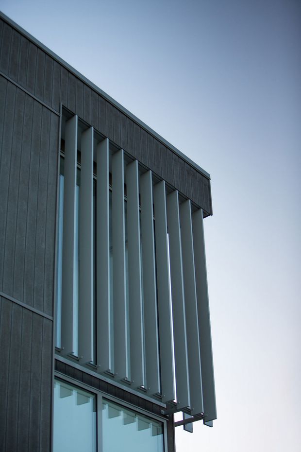 Close-up exterior architectural photograph of a contemporary building corner, featuring dark vertical cladding panels and a projecting row of tall pale metal vertical fins screening the glazing below. The composition emphasizes clean geometric lines, facade depth, and solar-shading elements against a clear sky.