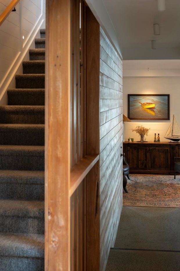 Interior view of a residential stairway and adjoining hallway or entry space. A dark carpeted staircase rises along the left beside a warm timber-framed balustrade or screen. The central wall is clad in horizontal pale timber boards, leading toward a furnished area with a dark wooden sideboard, framed coastal artwork, decorative objects, and a patterned rug. The space has a warm, refined mid-century-influenced character with restrained contemporary lighting.