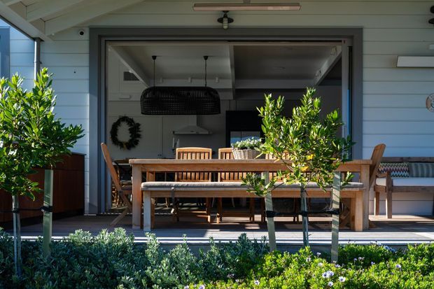 A sunny residential outdoor dining area on a timber deck, viewed from the garden. A long timber dining table with bench seating and chairs sits in front of large dark-framed sliding or folding glazing opening into a bright interior. The exterior has pale horizontal weatherboard cladding, a covered roof structure, potted and planted greenery, and a built-in bench along the wall. Interior kitchen and dining furnishings are visible through the open glazing.