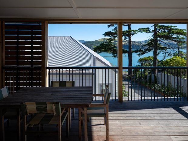 Covered outdoor dining terrace with timber decking, a large timber dining table, slatted privacy screening, and dark metal balustrades. The terrace overlooks a corrugated metal roof and a scenic coastal or lakeside landscape with hills and water visible beyond, framed by timber posts and glazing/openings.