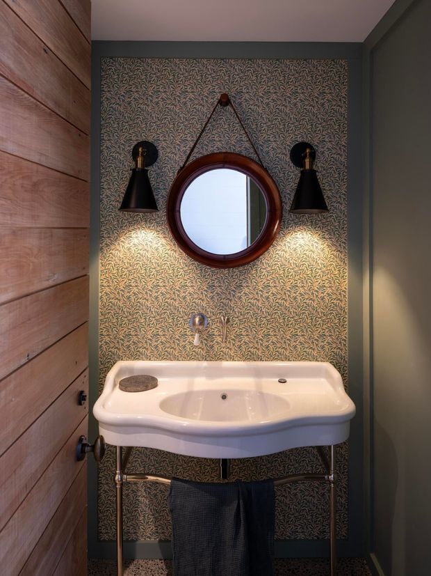 A compact, richly detailed bathroom washbasin vignette. A white vintage-style console basin with exposed polished metal legs is set against patterned botanical wallpaper framed in dark trim. A round dark timber-framed mirror hangs above the basin, flanked by two black wall sconces. Warm directional lighting highlights the wallpaper and basin, while a timber panelled door or wall is visible at left.