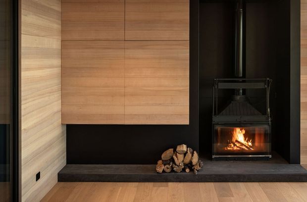 A refined contemporary fireplace feature wall with warm timber wall panelling and cabinetry above a dark stone hearth. A black wood-burning fireplace with a visible flame and stacked firewood creates a strong focal point, while timber flooring and dark wall surfaces add warmth and contrast.