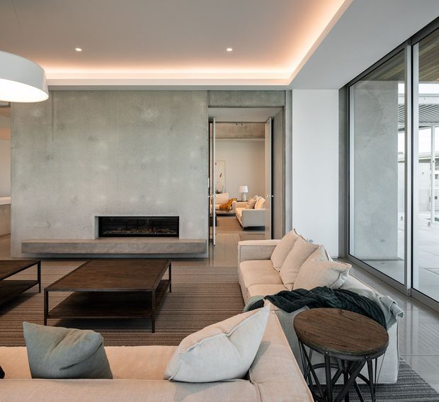 Contemporary living room with a large smooth concrete feature wall incorporating a linear fireplace and built-in hearth. A low sectional sofa, dark wood coffee tables, and a round side table occupy the foreground, while full-height black-framed glazing opens to a bright outdoor terrace. Recessed perimeter ceiling lighting adds a warm glow to the otherwise neutral, refined interior.