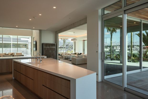Contemporary open-plan kitchen with a large wood-panelled island, pale stone benchtop, integrated sink and minimal cabinetry. Floor-to-ceiling glazed doors open to an outdoor terrace, with a bright living area and marina-like waterfront setting visible beyond. Recessed ceiling lighting and clean-lined finishes create an airy, refined interior.