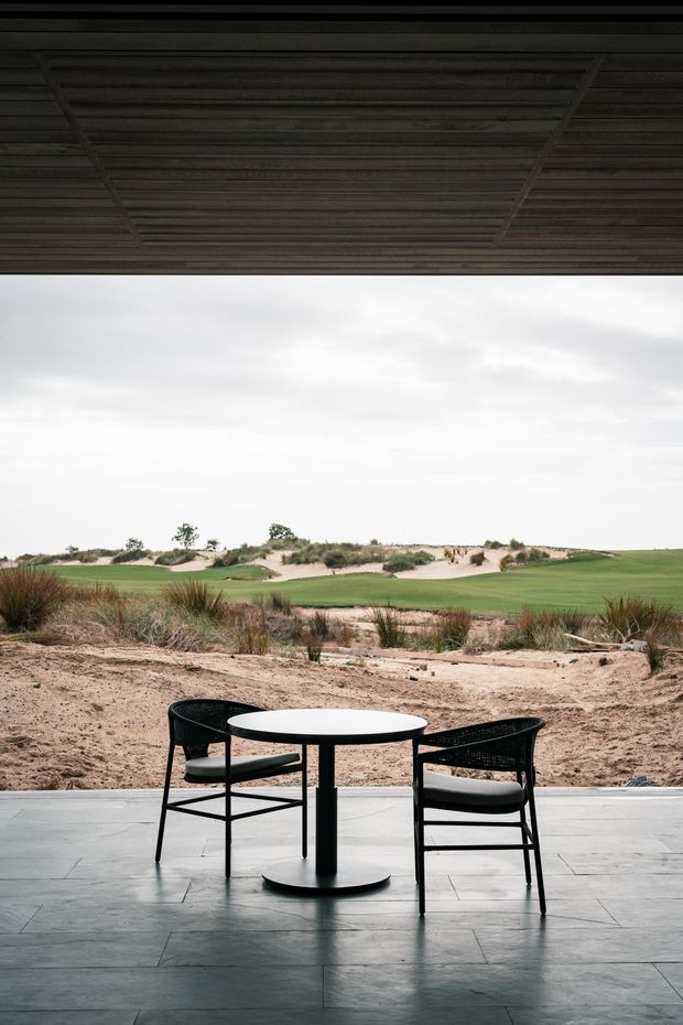 A minimalist outdoor dining setting beneath a dark timber-lined roof or covered terrace, with a round black pedestal table and two black woven chairs on large grey floor tiles. The open view looks across sandy coastal dunes and landscaped grass, creating a restrained connection between the sheltered space and the surrounding landscape.