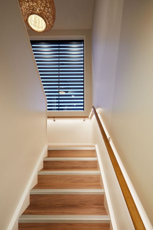 A narrow interior stairway with warm wood-finished treads, white painted walls and stringers, a timber handrail, and a woven pendant light. A tall window at the stair landing has horizontal roller blinds, bringing in soft daylight.