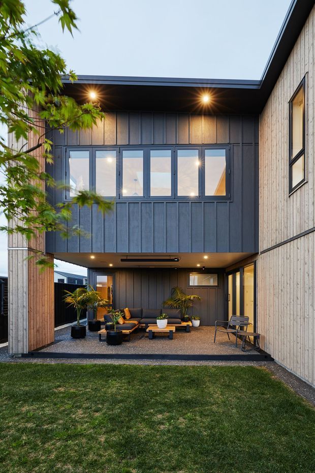 Exterior view of a contemporary two-storey home organized around a covered courtyard or outdoor lounge. The building features dark vertical cladding, pale timber-like vertical wall cladding, large aluminium-framed windows, a projecting upper-level volume, recessed ceiling lights, and a gravel-paved seating area with outdoor furniture and potted plants.