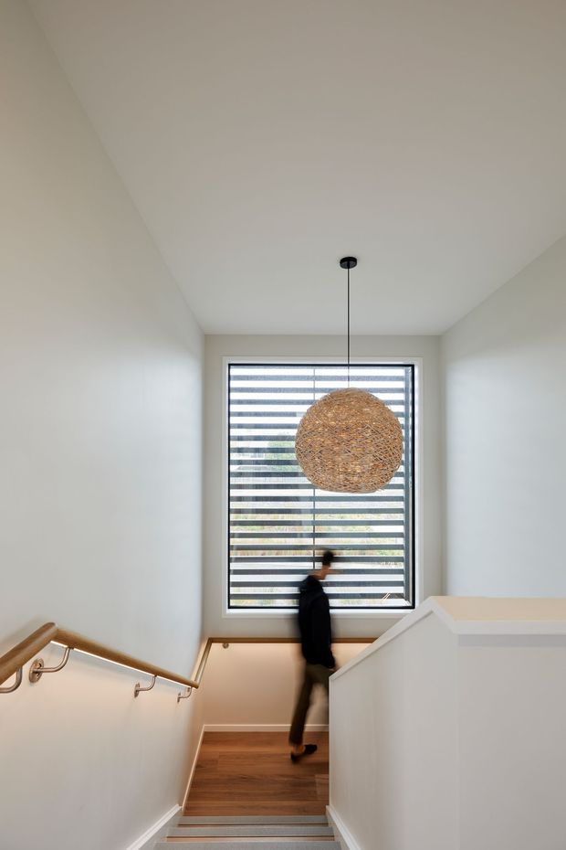 A bright, narrow stairway with white walls and ceiling, timber stair treads, a timber handrail with metal brackets, and a large woven spherical pendant light. A tall horizontal louvre window provides filtered natural light, while a person is blurred on the stairs.