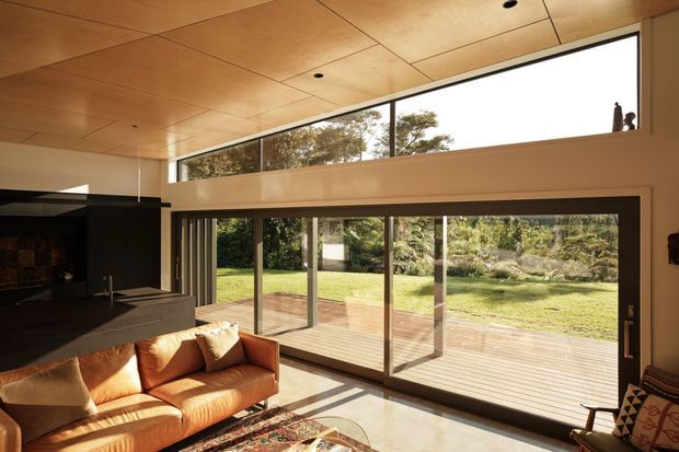 A warm, modern living room with a timber-panelled ceiling, polished concrete floor, and expansive floor-to-ceiling sliding glass doors opening onto a timber deck and landscaped garden. The room includes a large tan leather sofa, dark built-in cabinetry or fireplace surround, and strong indoor-outdoor connection. A high clerestory window and partial double-height volume add to the spacious feel.