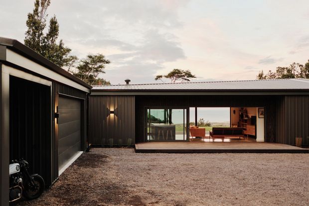 A low-slung contemporary single-storey residence viewed from a gravel courtyard or driveway. The dark vertically ribbed metal-clad building has a shallow pitched metal roof, an attached garage or outbuilding at left, and expansive sliding glass doors opening to a warmly lit living area. Interior furnishings and a kitchen island are visible through the glazing, with trees and an open landscape beyond.