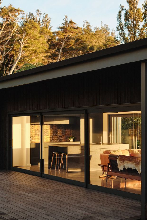 Exterior view of a modern single-storey home opening onto a broad timber deck. The dark, vertically clad facade has a strong sloping roofline and extensive black-framed glazing, revealing a warm open-plan kitchen and dining/living area inside. Bushland surrounds the building, with late-afternoon sunlight creating a warm, inviting atmosphere.