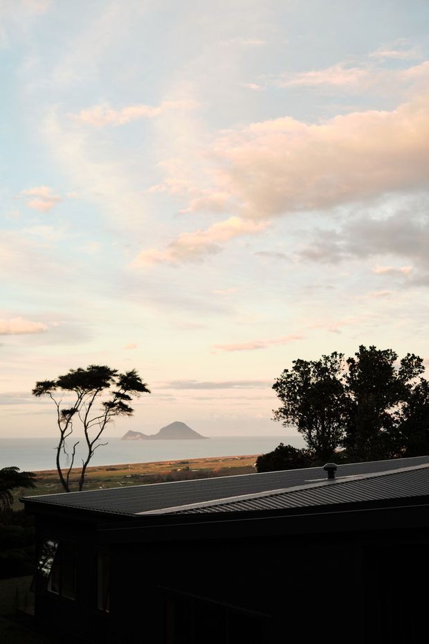 Exterior view of a low, dark-roofed building silhouetted against a coastal landscape at dusk, with the ocean, sandy shoreline, offshore island or headland, and windswept trees visible beyond. The image is dominated by pastel sky and expansive coastal scenery.