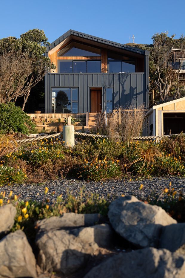 Exterior view of a compact contemporary coastal house set behind landscaped native planting and a rocky foreground. The two-storey facade combines dark vertical metal cladding, warm timber cladding beneath a low-pitched gable roof, large dark-framed glazed openings, and a timber entry stair. A separate small garage or outbuilding is visible at the right.