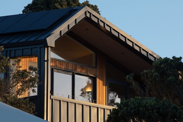 Exterior view of a contemporary, steeply pitched gable-roofed residence with a dramatic projecting roof canopy, dark metal roof and framing, warm vertical timber cladding, and large glazed openings. Dense landscaping partially screens the lower facade, while warm low-angle sunlight highlights the timber and recessed covered outdoor area.