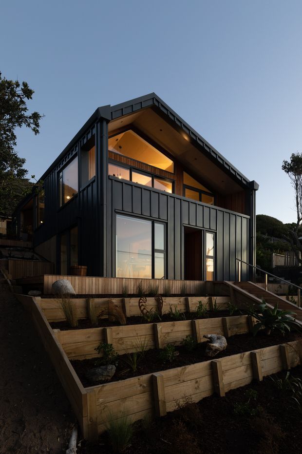 Exterior dusk photograph of a compact multi-level contemporary house on a sloped site. The home features dark vertical metal cladding, extensive glazing, a prominent steep gable roof with warm timber soffits, an upper balcony, and terraced timber retaining walls with planted garden beds in the foreground.