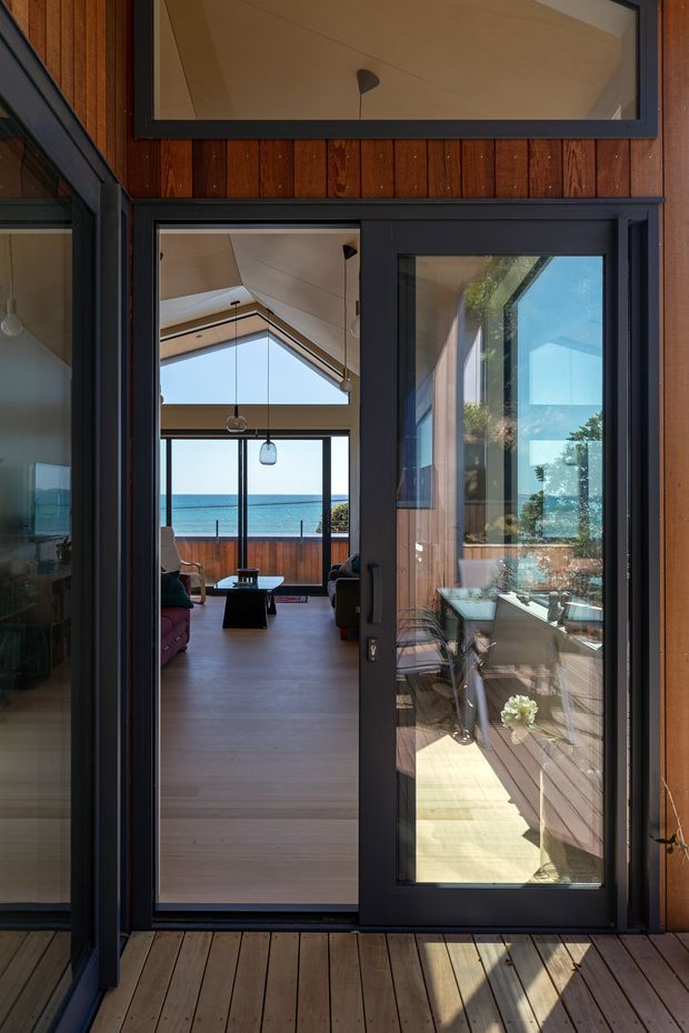 View from a timber deck through a dark-framed glazed doorway into a bright coastal living room. The interior has a high sloped ceiling, expansive glazing, pale timber flooring, pendant lights, and lounge furniture, with a clear ocean horizon visible beyond the far balcony. The exterior facade combines warm vertical timber cladding with charcoal window and door frames.