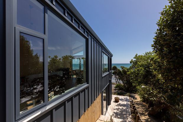 Exterior view of a contemporary dark-clad building on a coastal site, with large reflective windows, vertical facade detailing, a timber deck and glimpses of the ocean beyond dense vegetation.