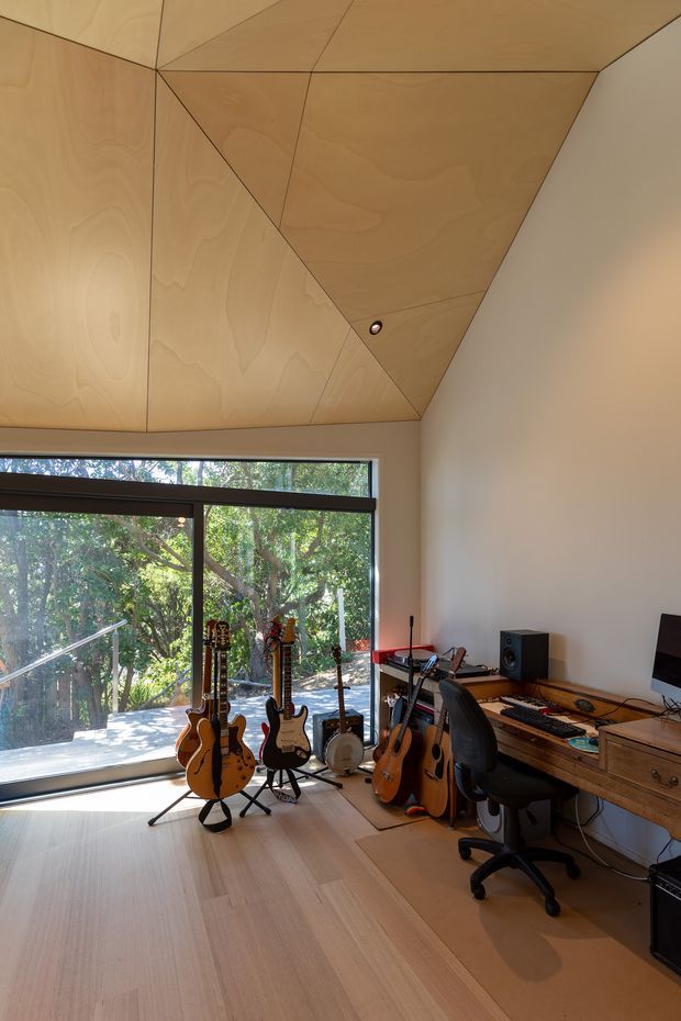 A compact music-focused home office or studio with several guitars and a banjo on stands, a desk with computer equipment and speakers, and extensive warm plywood paneling forming an angular geometric ceiling and upper wall surface. Pale timber flooring, a large dark-framed window, and views of leafy trees create a bright connection to the outdoors.