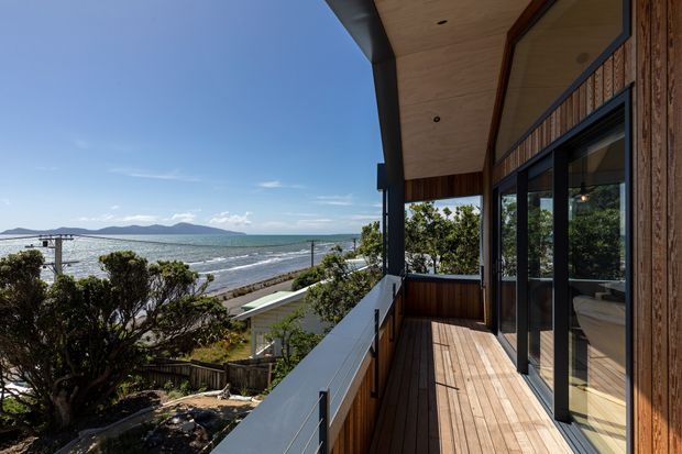 A contemporary coastal home balcony or covered deck overlooking a beach, ocean, and distant hills. The space features warm timber decking and soffit lining, dark metal structural posts and window frames, extensive glazing, and a simple white balustrade. The sheltered outdoor area connects directly to the glazed interior and has an open, elevated seaside outlook.