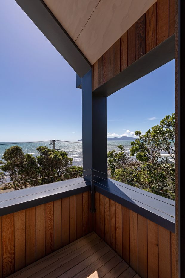 A compact elevated timber balcony or terrace overlooking a bright coastal bay, with dark painted structural posts and rails, warm vertical timber wall cladding, timber decking, and fine cable balustrades. The view includes shimmering water, a distant mountainous shoreline, and coastal vegetation. The image emphasizes the balcony’s corner construction and strong indoor-outdoor connection.