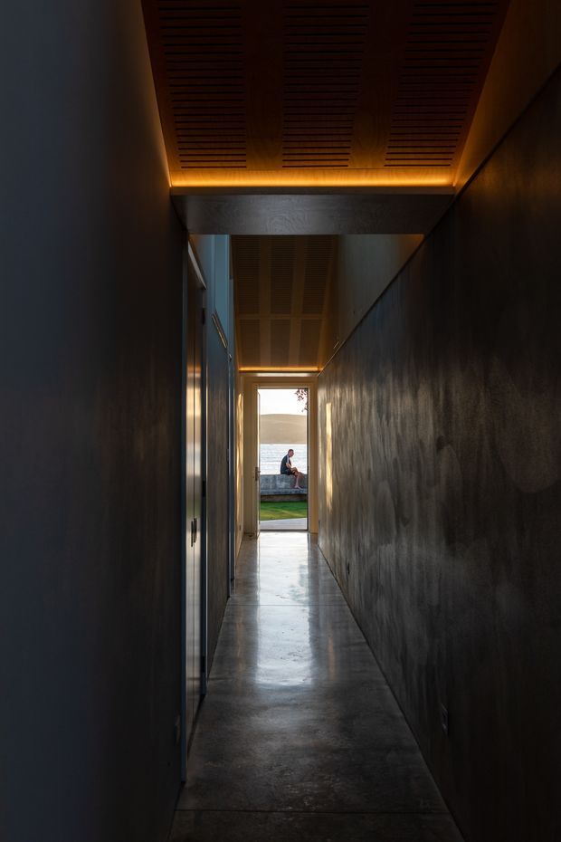 A narrow, elongated interior hallway with dark textured walls, a polished concrete floor, and a warm timber slatted ceiling with concealed linear lighting. Glazed or reflective openings line the left side, while the corridor terminates at an open doorway framing a small lawn, seated person, and distant landscape. The composition is moody and highly directional, emphasizing the length of the passage and contrast between dark interior finishes and bright exterior light.