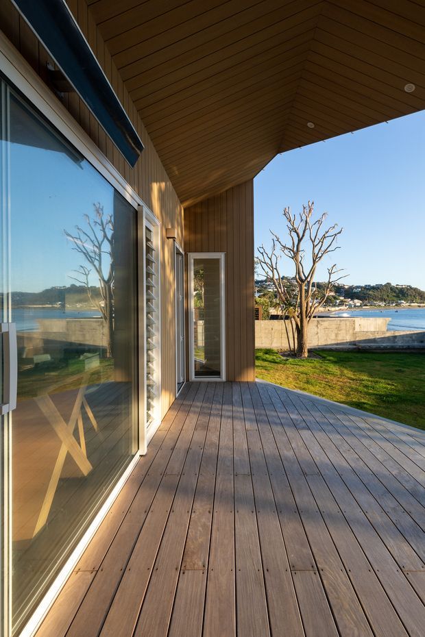 A covered timber deck or terrace beside a modern waterfront home. Wide glazed sliding doors reflect the interior, while vertical timber wall cladding and a richly lined timber soffit create a warm, contemporary architectural setting. The deck overlooks a lawn, retaining wall, calm water and the opposite shoreline.