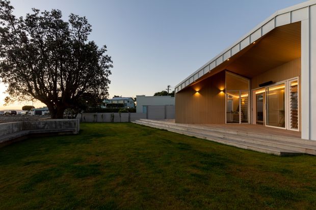 Exterior view of a low, modern single-storey building opening onto a broad lawn and paved terrace. The facade combines white metal-clad framing with warm vertical timber wall lining and a timber soffit beneath a deep overhanging roof. Full-height glazed doors and windows connect the interior to the terrace, with warm evening lighting visible inside and under the eaves. A mature tree and neighbouring low-rise buildings sit in the background.