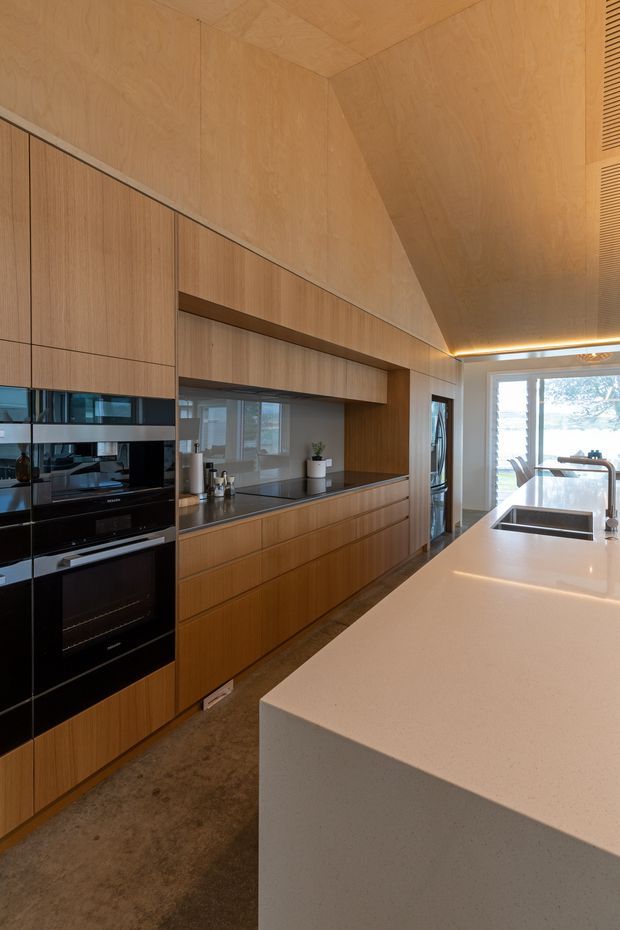 A warm, contemporary kitchen with extensive vertical-grain timber cabinetry and matching timber wall and ceiling lining. The room features integrated black appliances, a long dark benchtop, a large pale stone island with an undermount sink and tap, recessed open shelving, and concealed warm LED lighting along the ceiling. Glazing at the rear provides views outdoors.