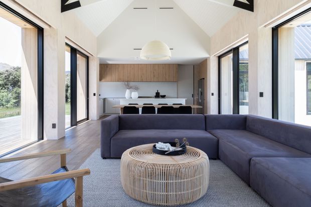 Bright contemporary open-plan living and dining space with a dramatic vaulted ceiling, extensive floor-to-ceiling glazing, pale timber wall finishes, and a minimalist kitchen at the rear. A large dark sectional sofa and woven round coffee table define the living area, while the dining table sits between the lounge and kitchen. The room opens visually to landscaped surroundings through wide glazed doors and windows.