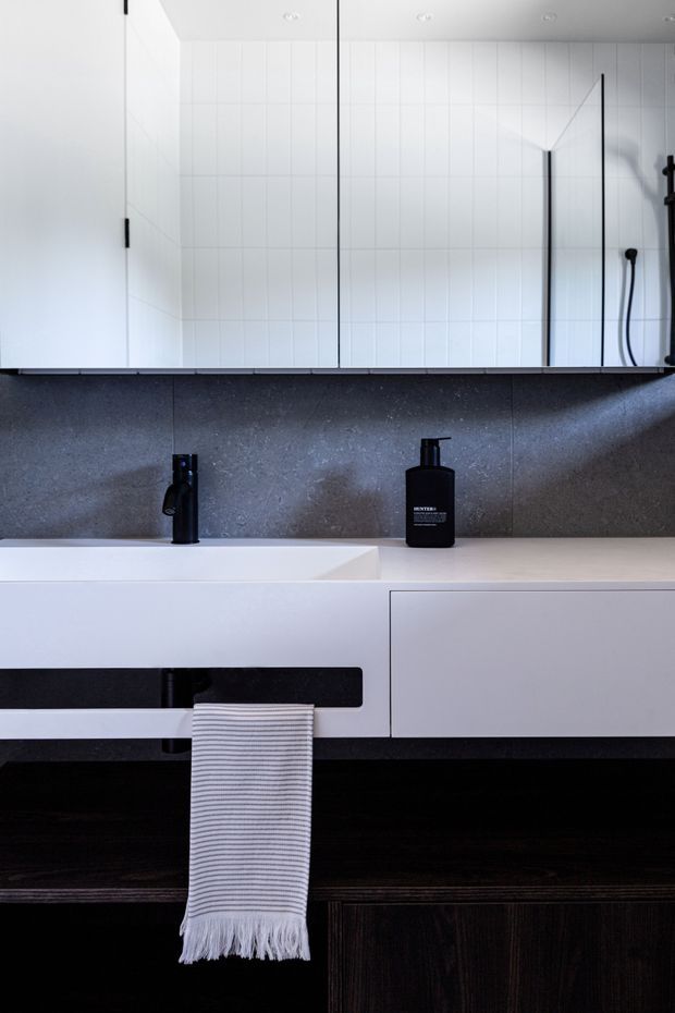 A contemporary minimalist bathroom vanity with a white integrated basin, wall-mounted white cabinetry, large mirrored cabinet, dark textured wall finish, black tapware and dark timber flooring or cabinetry. The space has clean linear detailing and a monochromatic black, white and grey palette.