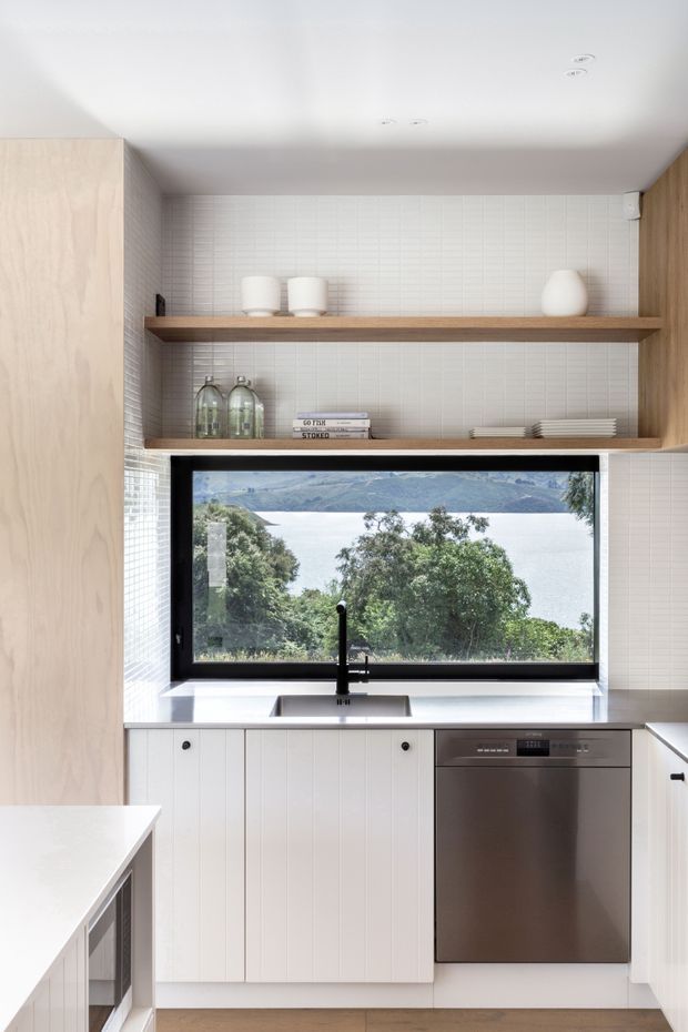 Bright contemporary kitchen with white vertical-panel cabinetry, a stainless-steel dishwasher, pale timber cabinetry and open shelving, and a white textured tiled splashback. A large black-framed window above the sink overlooks a lake and surrounding hills.