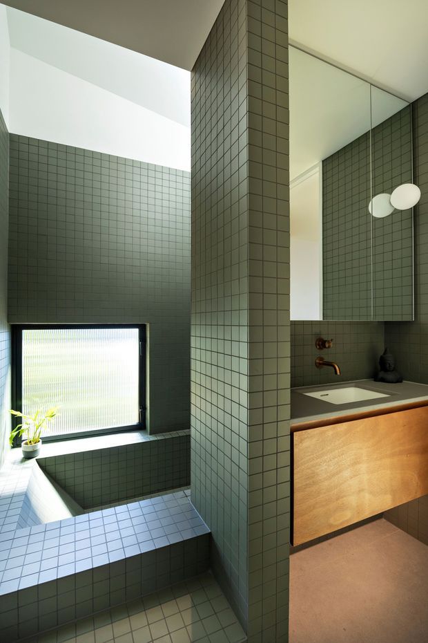 A contemporary bathroom with a deep green-gray small-format tiled bath area and full-height tiled dividing pier. The room includes a freestanding bathtub beneath a frosted window, a timber-front floating vanity with an integrated white basin, brushed bronze tapware, and a large mirrored cabinet. Natural light creates a calm, refined atmosphere.