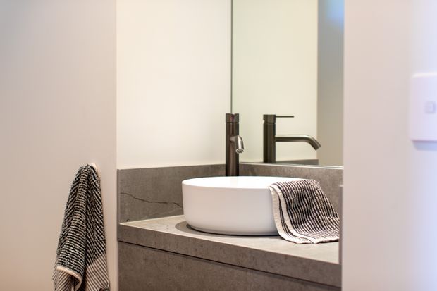 Close-up view of a contemporary bathroom vanity with a white vessel basin set on a grey stone benchtop, dark tapware, large mirror, and striped towels. The space has a clean, minimal, neutral-toned fit-out.