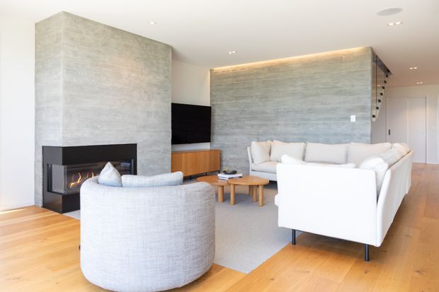 Bright contemporary living room with a large textured grey concrete-style feature wall and integrated fireplace, white sectional sofa, rounded upholstered armchair, timber flooring, low timber coffee tables, and concealed warm LED wall lighting. A television and timber media joinery sit beside the fireplace, with a hallway and stair area visible beyond.