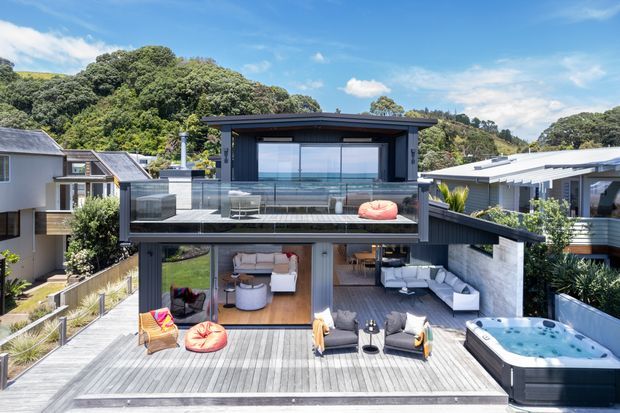 Exterior view of a contemporary multi-level home on a sloping, leafy site, featuring dark vertical cladding, extensive glazing, broad timber decking, a glass-balustraded upper terrace, outdoor lounge areas, and a spa pool. The design steps down the site with strong indoor-outdoor connections and elevated views.
