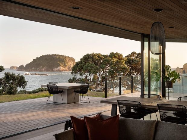 Open-plan coastal living and dining area extending onto a generous timber deck, with floor-to-ceiling glazing and a broad covered timber-lined ceiling. The space includes an outdoor dining setting, indoor dining table and upholstered seating, overlooking a rocky shoreline and vegetated island. The design has a refined contemporary indoor-outdoor connection and expansive coastal views.