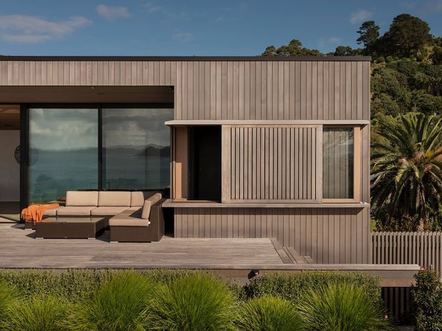 Exterior view of a contemporary, flat-roofed residential building clad in vertical timber boards. Large dark-framed glazing opens onto a timber deck with outdoor sectional seating, while sliding timber screening and a raised lower level create a layered facade. Dense planting and a palm tree surround the home, with a coastal landscape visible through the glazing.