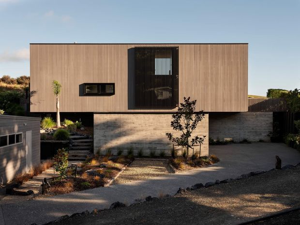 Street-facing exterior view of a contemporary two-level residence. The upper volume has a long, finely ribbed timber-toned facade with a dramatic dark vertical screen enclosing a recessed window or balcony. A pale textured masonry base supports the upper storey, while a planted gravel forecourt, steps, driveway and boundary elements frame the home.