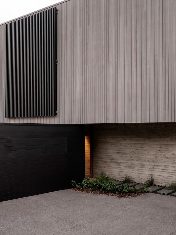 A close-up view of a contemporary residential facade showing pale vertical cladding, a dark vertical privacy screen, a black sectional garage door, and a textured concrete block wall. A narrow warm-lit entry recess and a small planted garden bed sit beside the driveway, with grey concrete paving in the foreground.