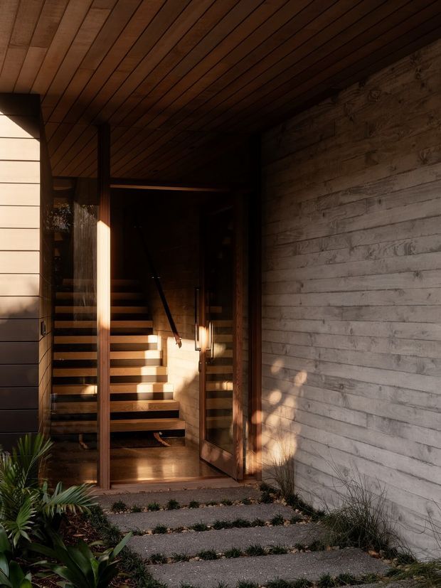 A dramatic residential entrance viewed from an outdoor garden path. Broad concrete stepping slabs lead to a timber-framed glazed entry door, opening onto a warmly lit stairway. The entry features extensive horizontal timber wall and ceiling lining, a rough pale masonry or concrete-block side wall, timber stairs with dark metal handrail, and integrated warm lighting. Strong afternoon sunlight creates deep shadows and a warm, secluded atmosphere, with low planting along the path.