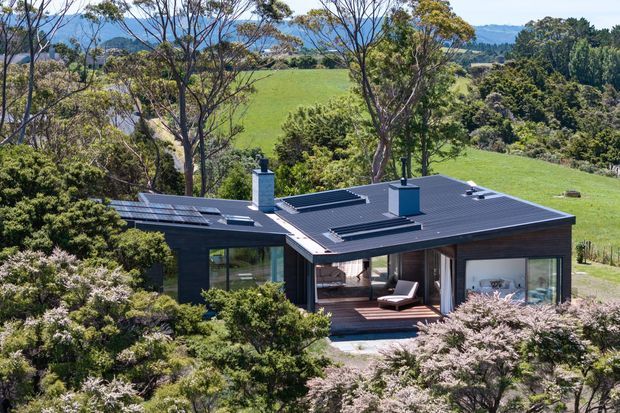Aerial exterior view of a low, modern single-storey house set within a lush rural landscape. The home has a dark standing-seam metal flat or very low-pitched roof, dark cladding, extensive glazing, multiple roof skylights or vents, and a central outdoor terrace with timber decking. Rolling green farmland and mature vegetation surround the property.