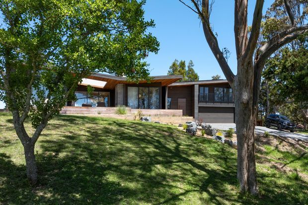 Exterior view of a low, single-storey modern residence set within a mature landscaped garden. The house has a long horizontal composition, timber-lined projecting roof forms, extensive glazing, dark vertical wall panels, a timber-clad entry volume, and an integrated garage. A gently sloping lawn, established trees, stone edging and driveway frame the building.