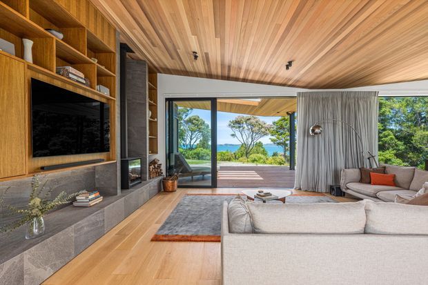 A warm, sophisticated living room with extensive timber lining across the ceiling and built-in entertainment wall. The room features a large pale sectional sofa, grey area rug, stone fireplace and hearth, timber flooring, open shelving, floor-to-ceiling glazing, and sheer curtains opening onto a deck with a distant coastal view.