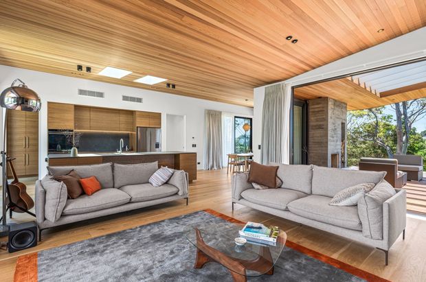 Open-plan living room with two large grey sofas, a glass and timber coffee table, and a grey area rug. The room features extensive warm timber lining across the ceiling, timber flooring, and a built-in kitchen with dark timber cabinetry and a reflective splashback. Full-height glazed openings connect the living area to a timber-covered outdoor deck, while a stone-clad fireplace or column is visible near the opening. Contemporary furnishings and abundant daylight create a refined, warm interior.